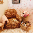 A cozy miniature-style room scene featuring a small floral upholstered armchair with a cream knit throw blanket, a round white crocheted rug, a woven basket with magazines, and a vase of pink roses against floral wallpaper.