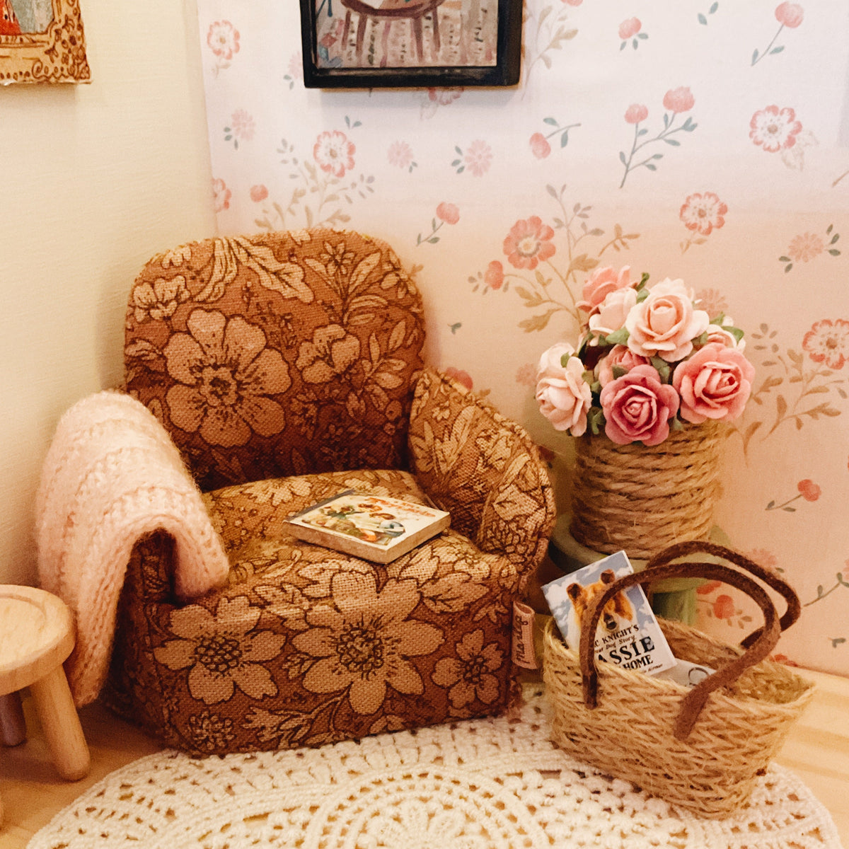 A cozy miniature-style room scene featuring a small floral upholstered armchair with a cream knit throw blanket, a round white crocheted rug, a woven basket with magazines, and a vase of pink roses against floral wallpaper.