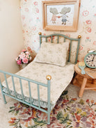 A Maileg miniature dollhouse-style single bed with a light blue metal frame and gold ball finials, dressed in cream bedding with a floral print. The bed is styled in a vintage room setting with matching floral wallpaper, a small wooden side table with a clock, and a framed picture of two Maileg mice on the wall.