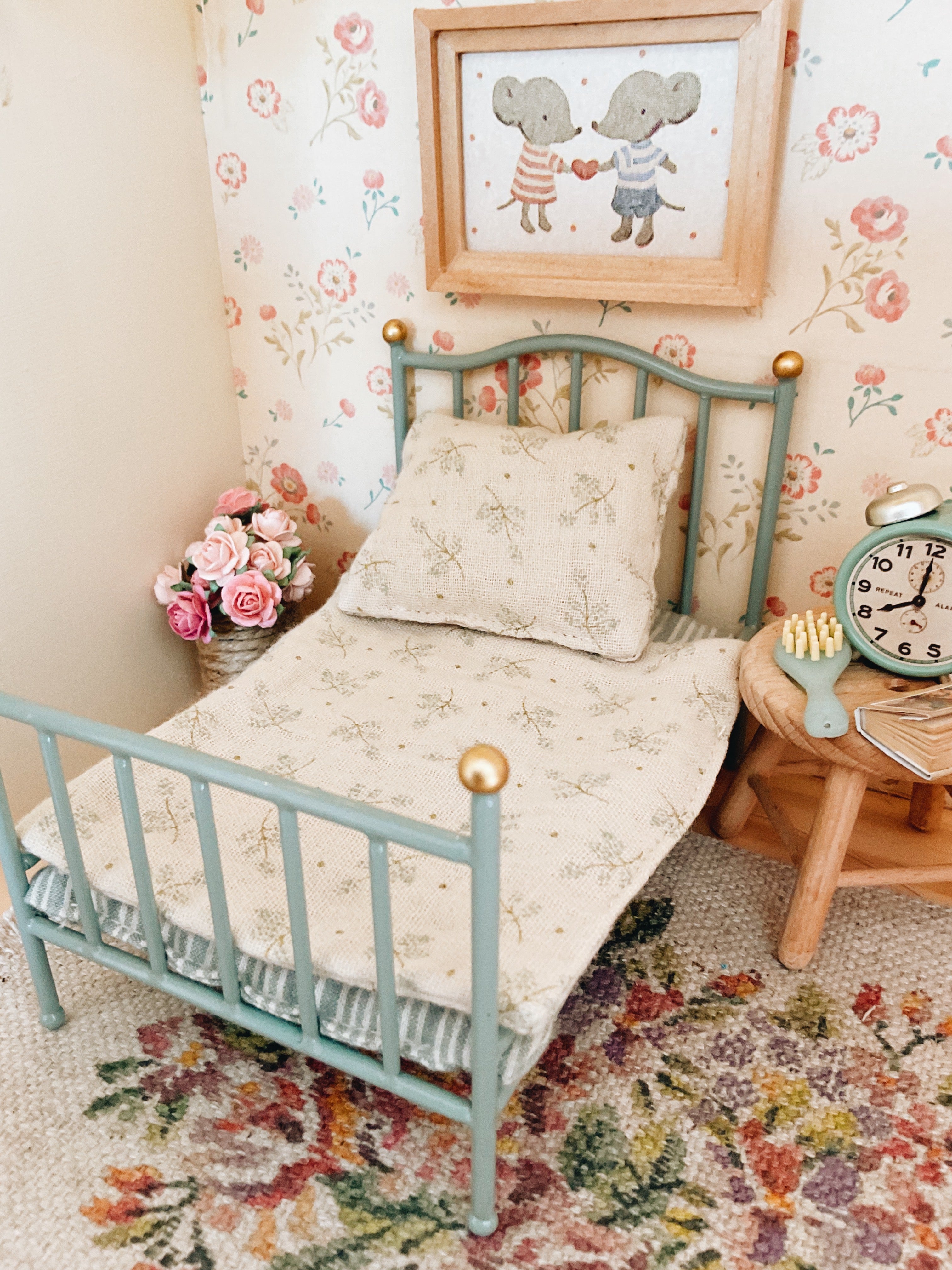 A Maileg miniature dollhouse-style single bed with a light blue metal frame and gold ball finials, dressed in cream bedding with a floral print. The bed is styled in a vintage room setting with matching floral wallpaper, a small wooden side table with a clock, and a framed picture of two Maileg mice on the wall.
