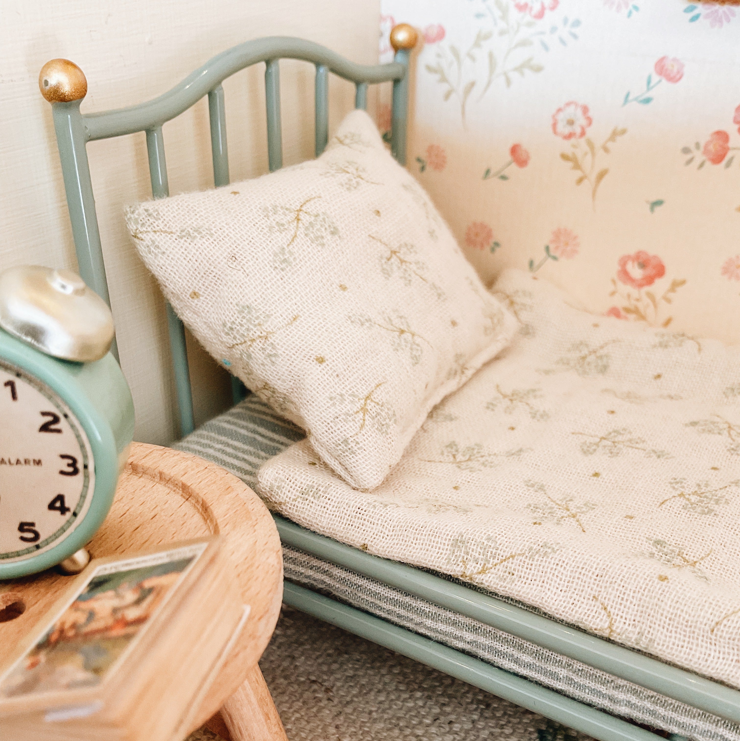 A maileg miniature dollhouse bed with a sage green frame, cream floral bedding, and a matching pillow, styled next to a small wooden side table and a matching miniature alarm clock.