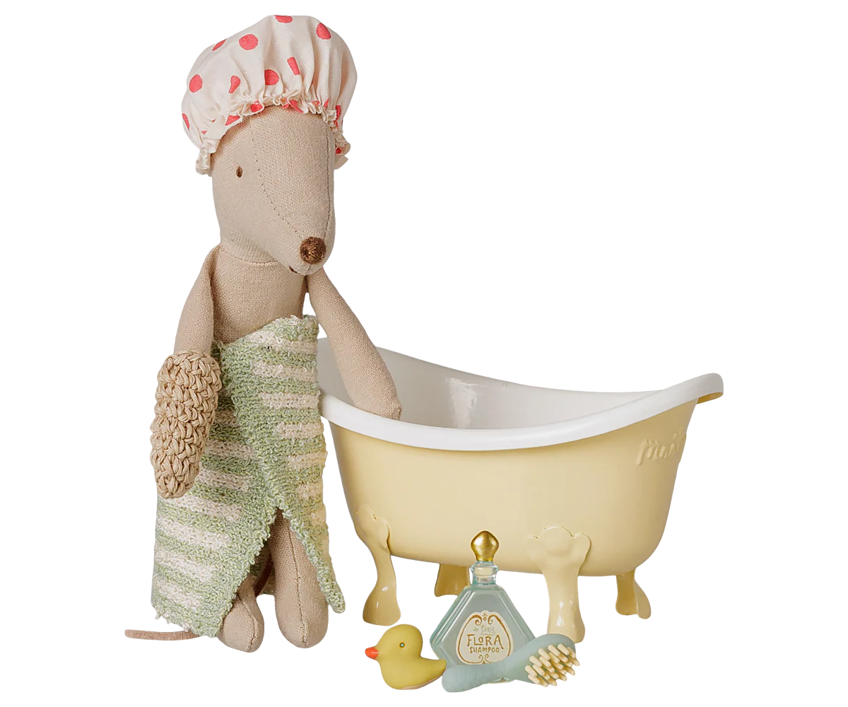 A toy bath scene featuring a beige plush mouse wearing a pink polka-dot shower cap and a green striped towel, sitting beside a cream-colored clawfoot bathtub. A small yellow rubber duck, a blue bottle, and a brush are placed in front on a white background.