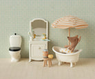 Miniature bathroom set with bathtub, toilet, and teddy bear under an umbrella against a patterned wall.
