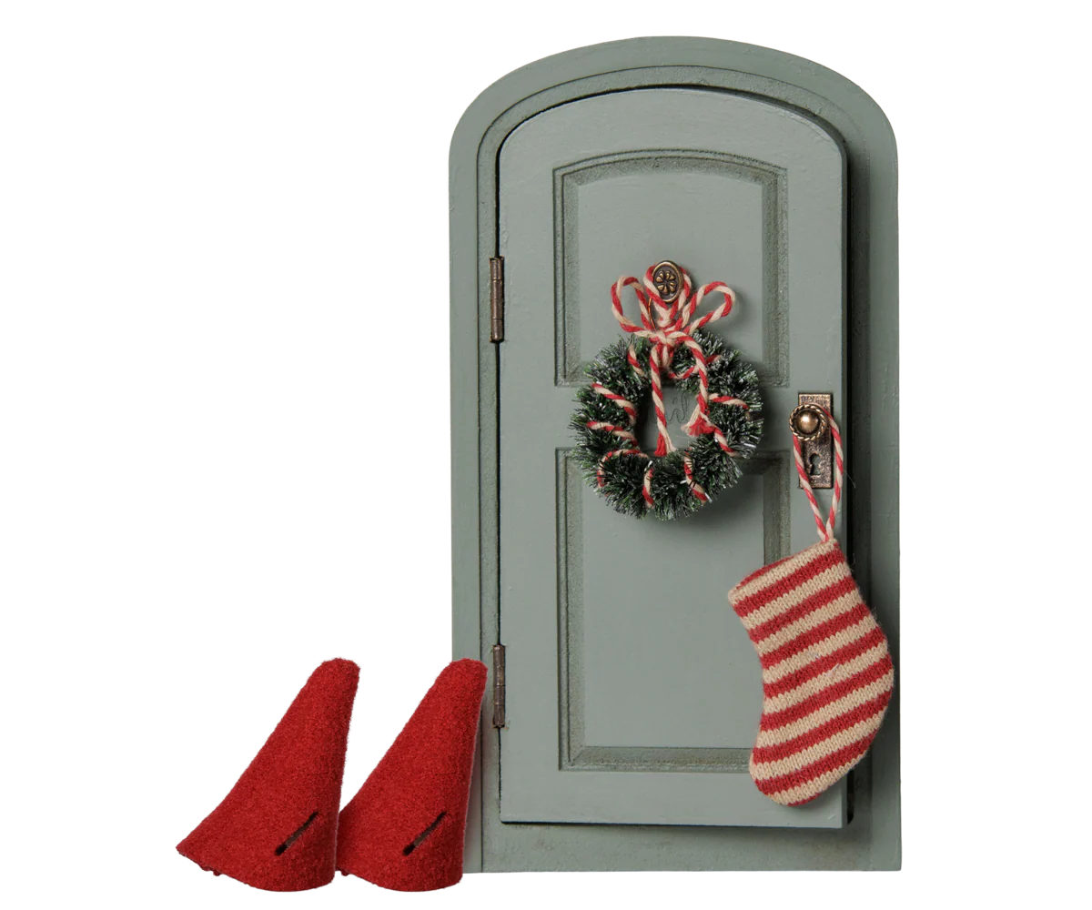 A miniature dollhouse-style gray door decorated with a green wreath, candy cane, and a red-and-white striped stocking, with two small red boot shapes beside it on a white background.