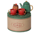 A decorative tea set displayed on a round tin canister. The set includes a green teapot with gold trim, two matching red teacups with saucers, and a small brown pumpkin decoration.