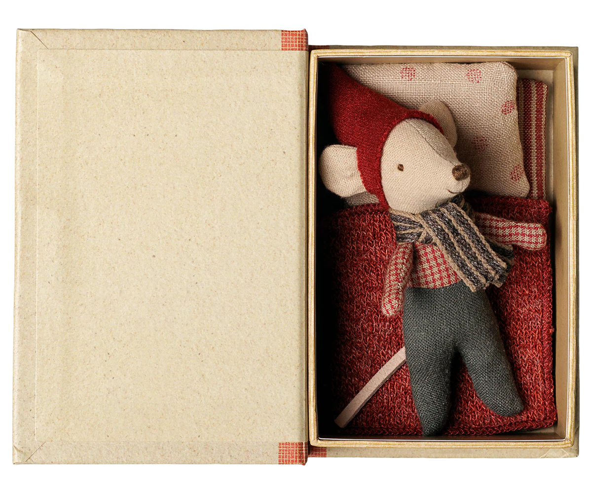 A small plush mouse toy dressed in a red hat and scarf with gray pants, presented inside an open beige gift box with a red patterned cushion.