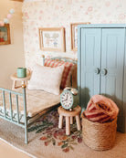 A styled miniature Maileg dollhouse bedroom scene featuring a light blue vintage bed with a patterned quilt and pillow, a matching blue closet, a small wooden side table with a mint green mug, a vintage-style green alarm clock on a stool, and a woven basket holding a pink knit blanket.
