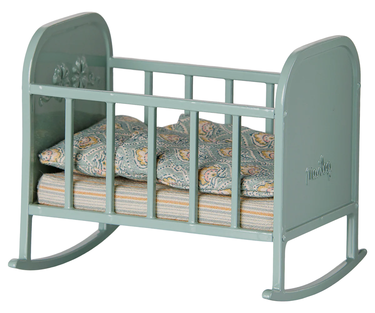 A miniature dollhouse rocking bed in a muted sage green color, furnished with a patterned blue and orange paisley quilt and a striped mattress, shown against a plain white background.