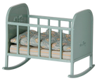 A miniature dollhouse rocking bed in a muted sage green color, furnished with a patterned blue and orange paisley quilt and a striped mattress, shown against a plain white background.
