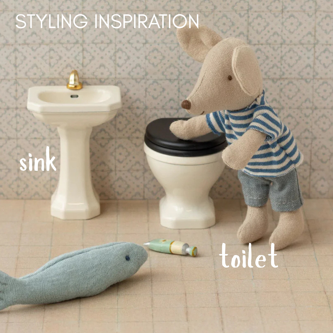 Toy maileg mouse in striped shirt and shorts standing next to toy sink and toilet with 'sink' and 'toilet' labels.