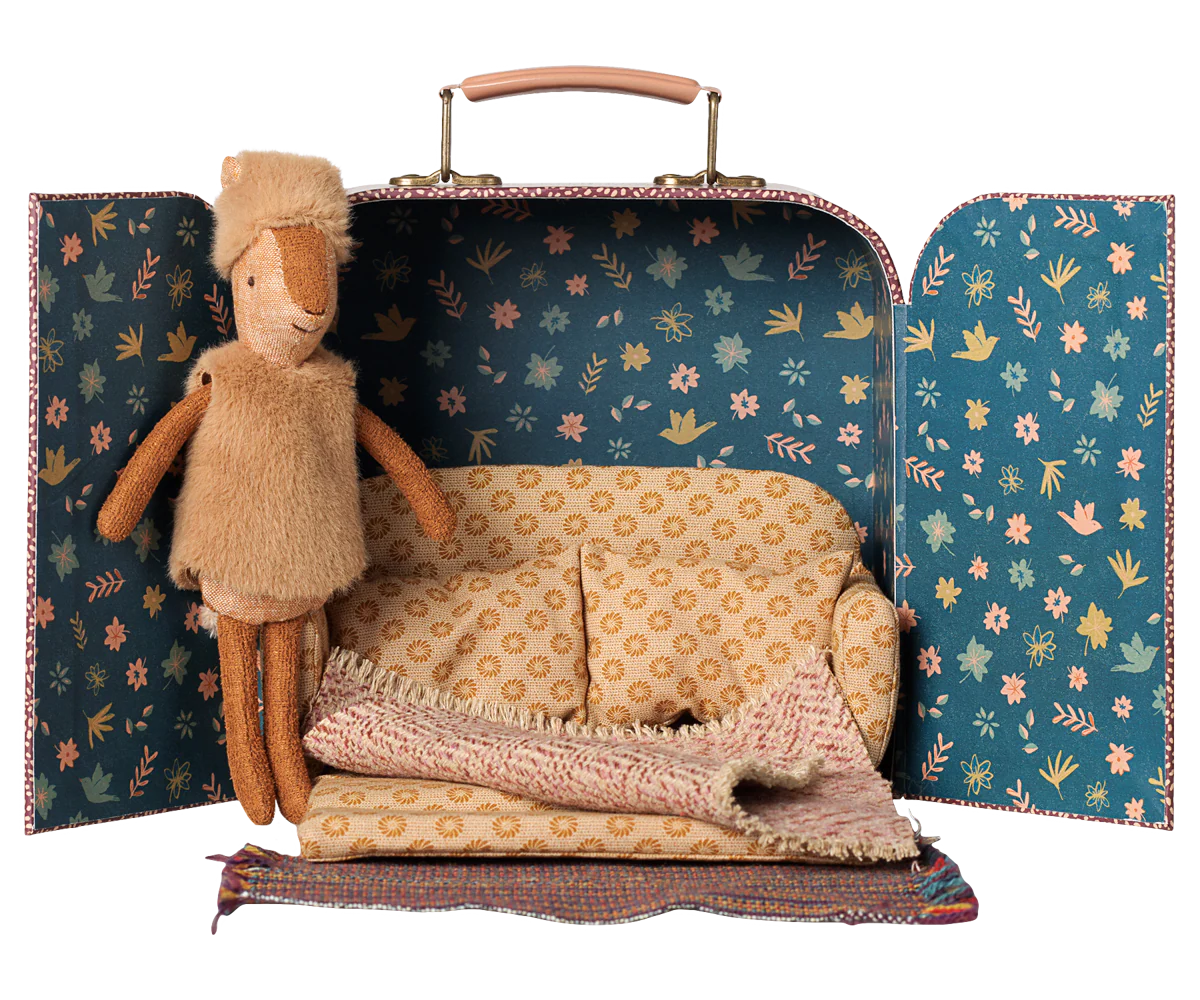 A miniature doll playset inside an open vintage-style suitcase, featuring a plush tan doll, a tiny patterned sofa, a knitted blanket, and a small rug on a white background.