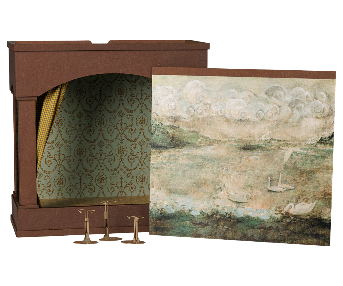 A decorative fireplace mantel display set featuring a brown faux-brick fireplace insert with patterned green and gold damask wallpaper lining, a matching landscape art print depicting swans on water, and three brass candlesticks.