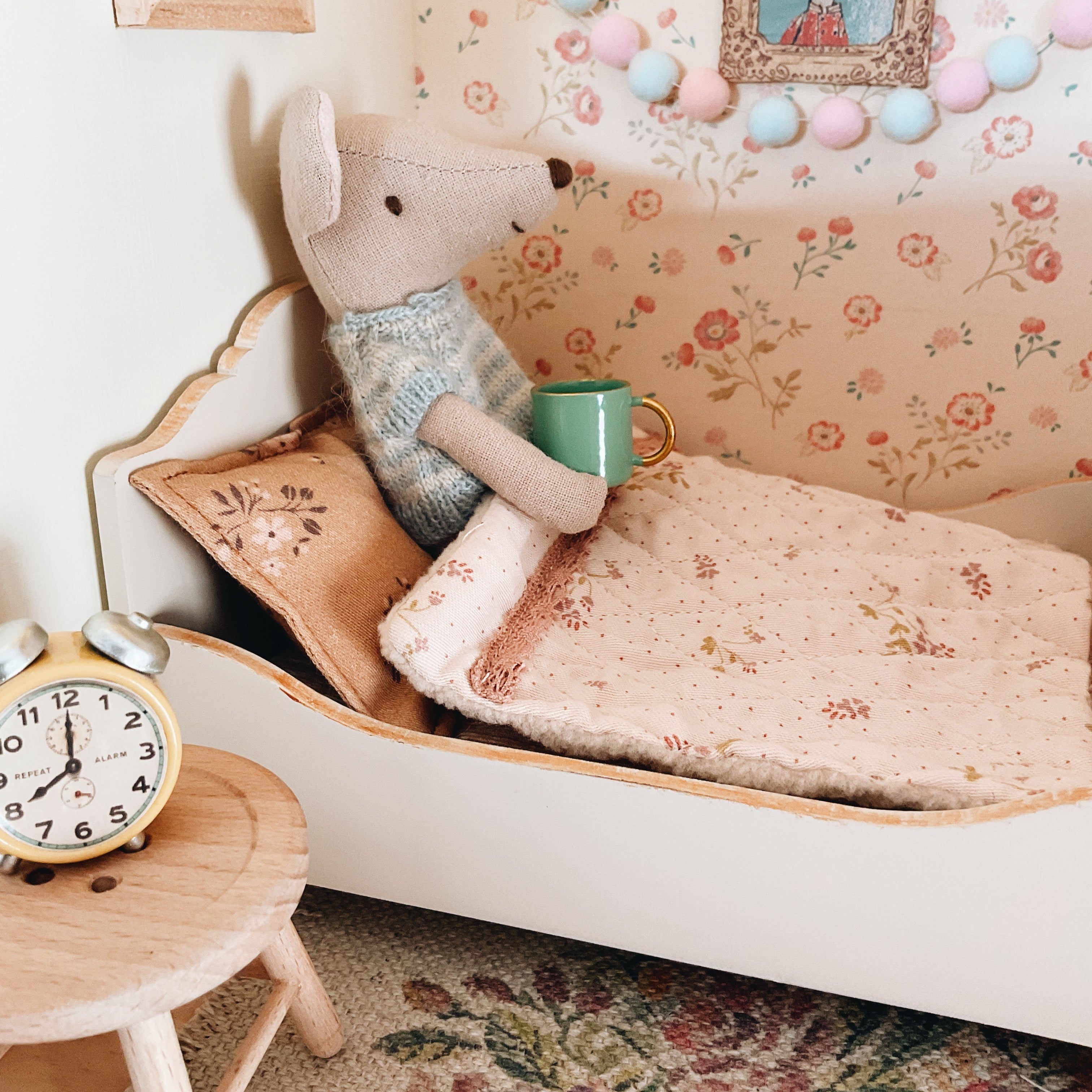 Maileg Small stuffed mouse with a mug on a floral-patterned bed in a cozy room.