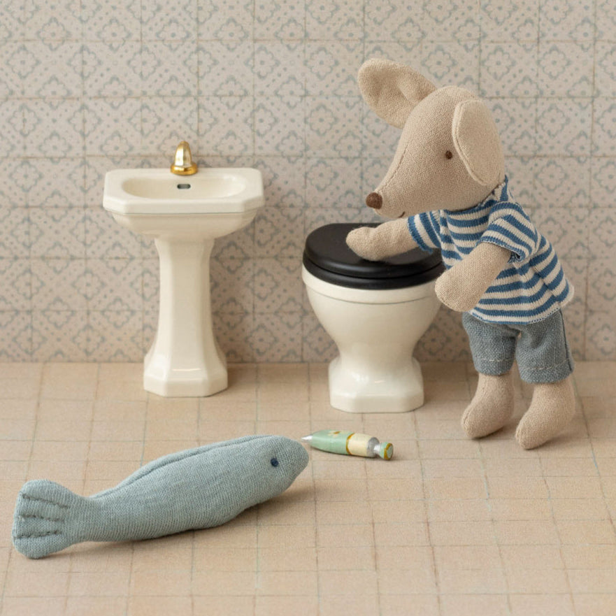Miniature Maileg mouse character using a toy toilet with a fish  nearby, set against a tiled bathroom wall.
