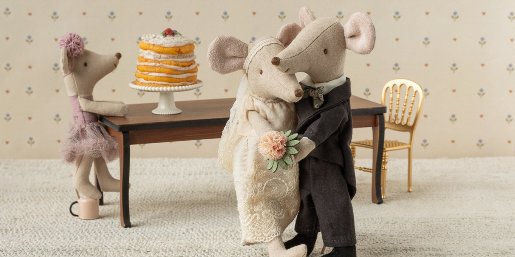 Maileg wedding couple holding hands dressed in wedding outfits in front of a big table with a wedding cake on top with a ballerina mouse trying to reach the cake 