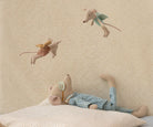 Stuffed Maileg maxi slze mouse in pyjamas lying on a bed with decorative wallpaper in the background and two maileg fairies flying above 