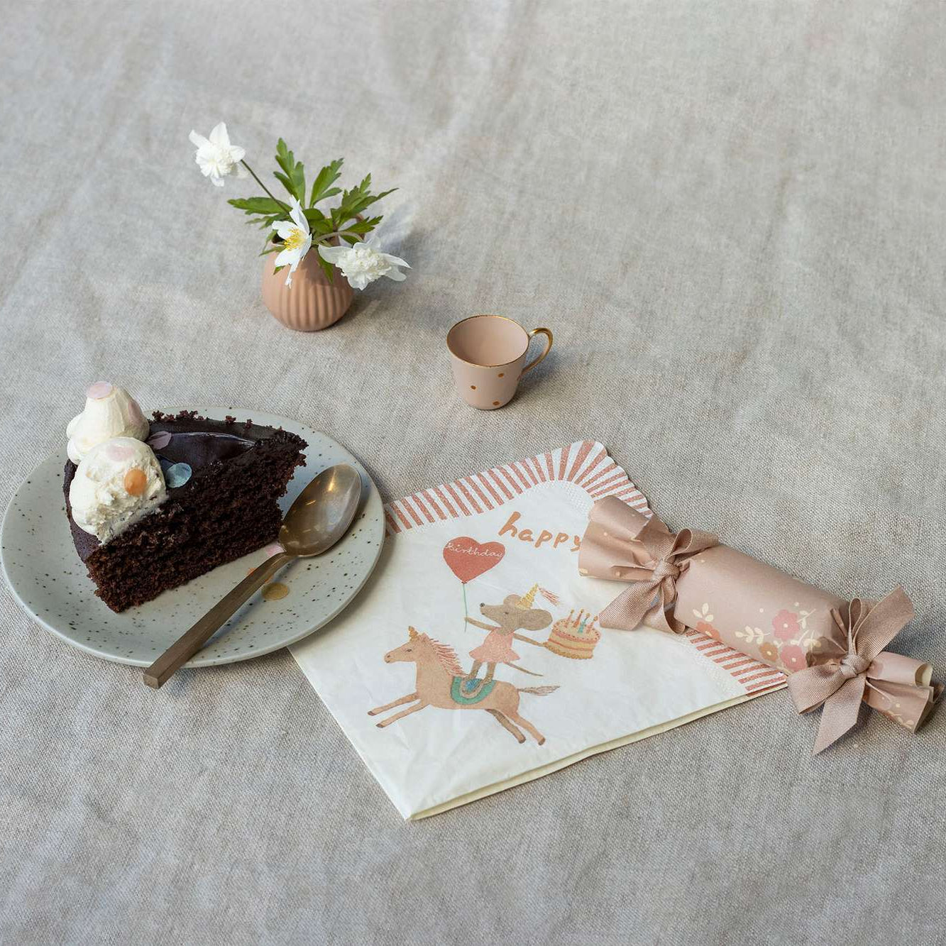 Chocolate cake with white frosting on a plate, accompanied by a mug, flowers, and a wrapped gift on a textured surface.