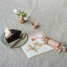 Chocolate cake with white frosting on a plate, accompanied by a mug, flowers, and a wrapped gift on a textured surface.