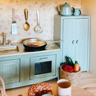 A styled miniature play kitchen set in a dollhouse scale, featuring a mint green toy stove and cabinet, a frying pan with toy food, and a small basket of toy vegetables, set against a floral wallpaper background.