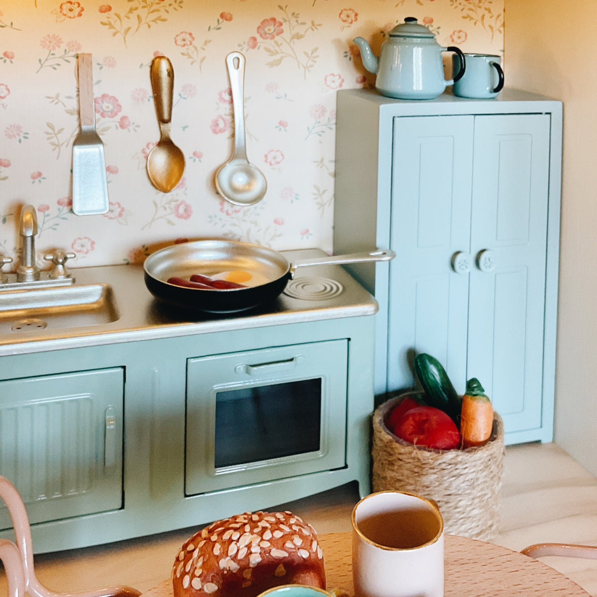 A styled miniature play kitchen set in a dollhouse scale, featuring a mint green toy stove and cabinet, a frying pan with toy food, and a small basket of toy vegetables, set against a floral wallpaper background.