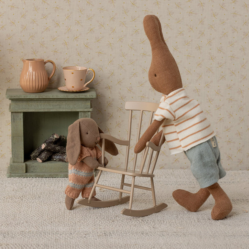 Two toy bunnies in a room with a lamp, small table, and rocking chair.