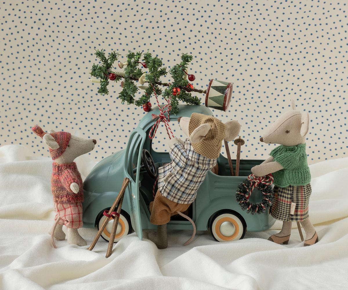 Three plush toy mice dressed in winter clothing gather around a small teal toy truck on white fabric, with one mouse loading a decorated pine branch into the truck bed. The background is a cream fabric with small blue polka dots.