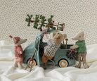 Three plush toy mice dressed in winter clothing gather around a small teal toy truck on white fabric, with one mouse loading a decorated pine branch into the truck bed. The background is a cream fabric with small blue polka dots.