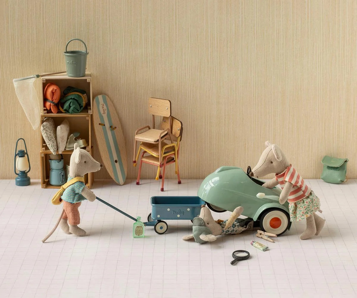A styled miniature scene with small animal figurines and toy props arranged on a white floor against a beige wall. One figurine pushes a blue wagon while another stands beside a mint green toy car, with additional beach-themed accessories and furniture in the background.