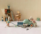 A styled miniature scene with small animal figurines and toy props arranged on a white floor against a beige wall. One figurine pushes a blue wagon while another stands beside a mint green toy car, with additional beach-themed accessories and furniture in the background.