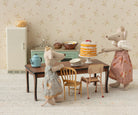 maileg Two toy mice in dresses standing at a table with a cake and tea set, in front of a floral wallpapered wall.