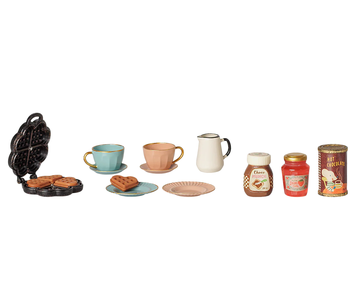 A miniature toy breakfast set featuring a black waffle iron with cookies, a light blue cup and saucer, a peach cup and saucer, a white pitcher, a blue plate with a cookie, a peach plate, two jars of jam, and a can of coffee, arranged on a plain white background.