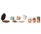 A miniature toy breakfast set featuring a black waffle iron with cookies, a light blue cup and saucer, a peach cup and saucer, a white pitcher, a blue plate with a cookie, a peach plate, two jars of jam, and a can of coffee, arranged on a plain white background.