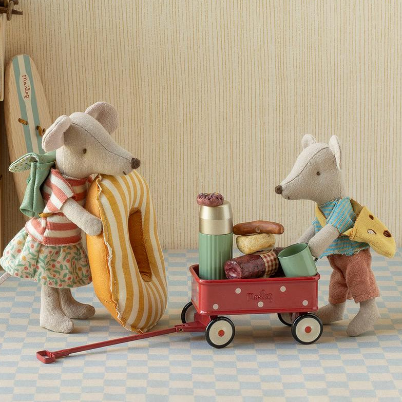A styled indoor scene with two plush mouse figures dressed in picnic clothing, one carrying a striped donut and the other pushing a red wagon loaded with picnic items, beside a wooden shelf with camping accessories and a lantern.