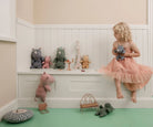 Child in a pink dress sitting on a white shelf with stuffed animals in a room with green floor and beige walls.
