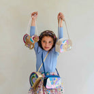 A young girl with curly brown hair wearing a light blue long-sleeve shirt and a colorful patterned skirt holds up three pastel unicorn and rainbow crossbody bags against a plain off-white wall.