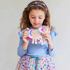 A young girl with curly brown hair wearing a light blue long-sleeve top and a colorful skirt, holding a handmade felt unicorn craft. She is wearing a matching felt headband against a plain light gray background.
