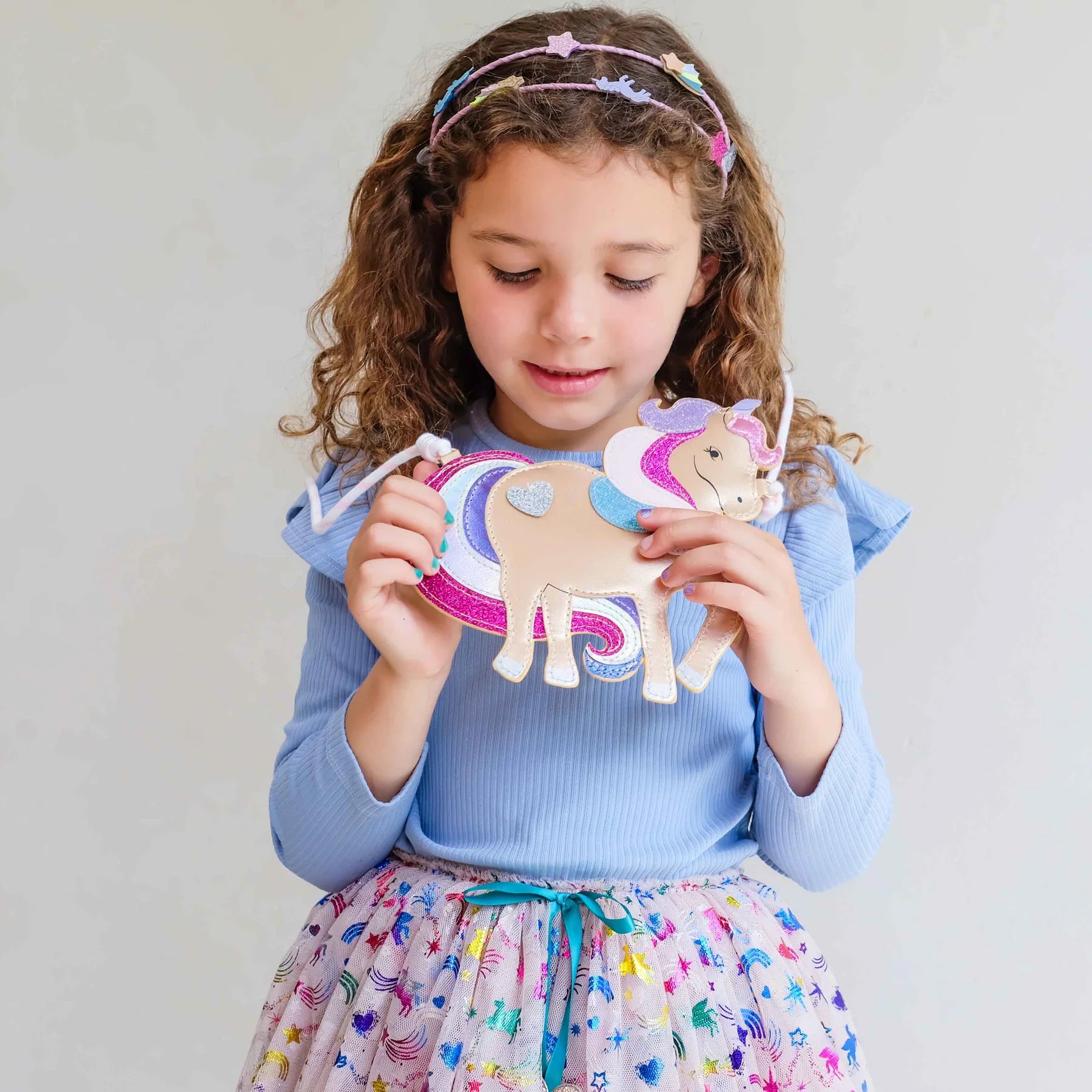 A young girl with curly brown hair wearing a light blue long-sleeve top and a colorful skirt, holding a handmade felt unicorn craft. She is wearing a matching felt headband against a plain light gray background.