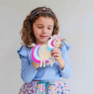 A young girl with curly brown hair wearing a light blue top and a colorful skirt, holding a wooden unicorn toy with glittery pink and blue details against a plain light gray background.