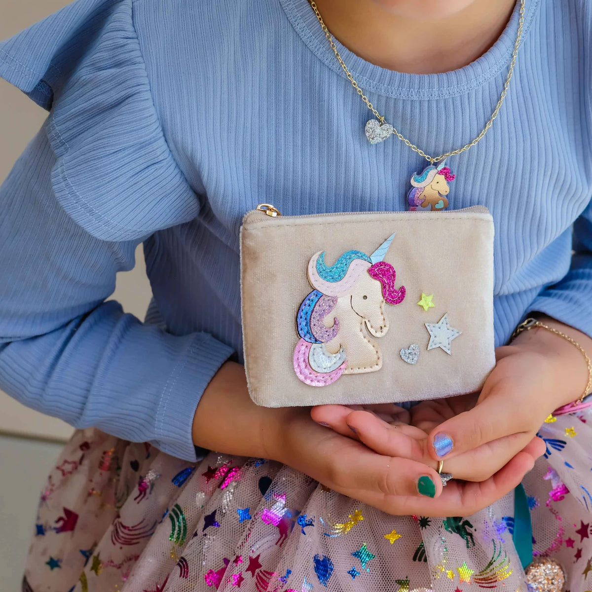 A child wearing a light blue ribbed long-sleeve top and a sparkly tulle skirt holds a small beige velvet pouch decorated with a colorful embroidered unicorn and star details.