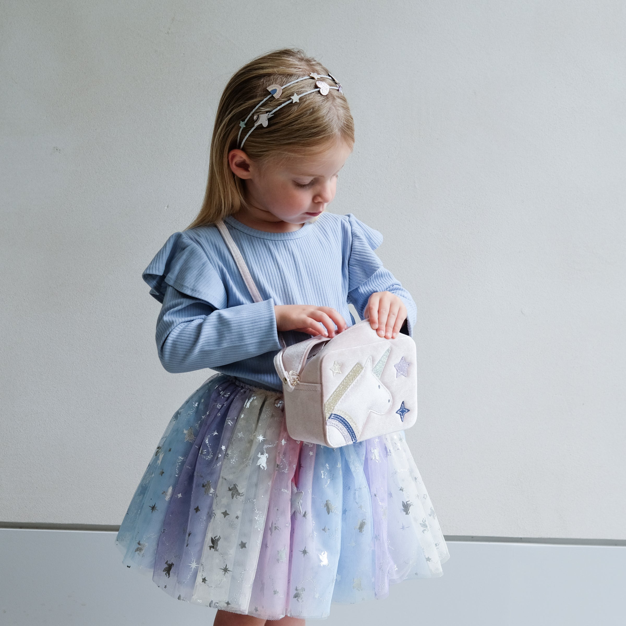 Young girl in a light blue sweater and star-patterned skirt holding a unicorn bag with a white background.