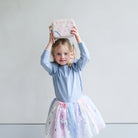 Young girl in a light blue top and colourful tutu holding a pink unicorn bag above her head against a plain background.