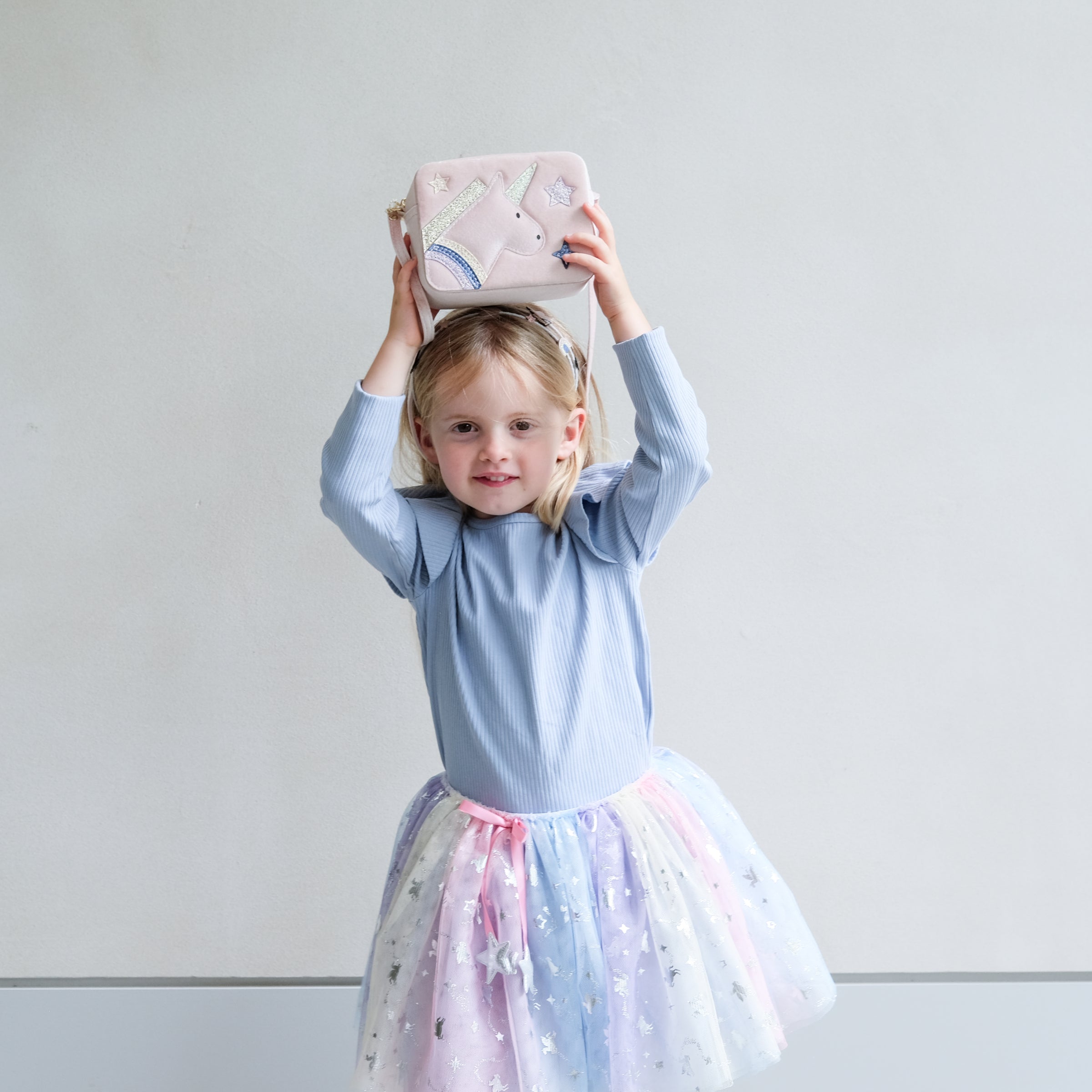 Young girl in a light blue top and colourful tutu holding a pink unicorn bag above her head against a plain background.