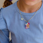 A close-up of a child wearing a gold chain necklace with a colorful unicorn pendant and a small glittery heart charm, paired with a light blue ribbed long-sleeve top.
