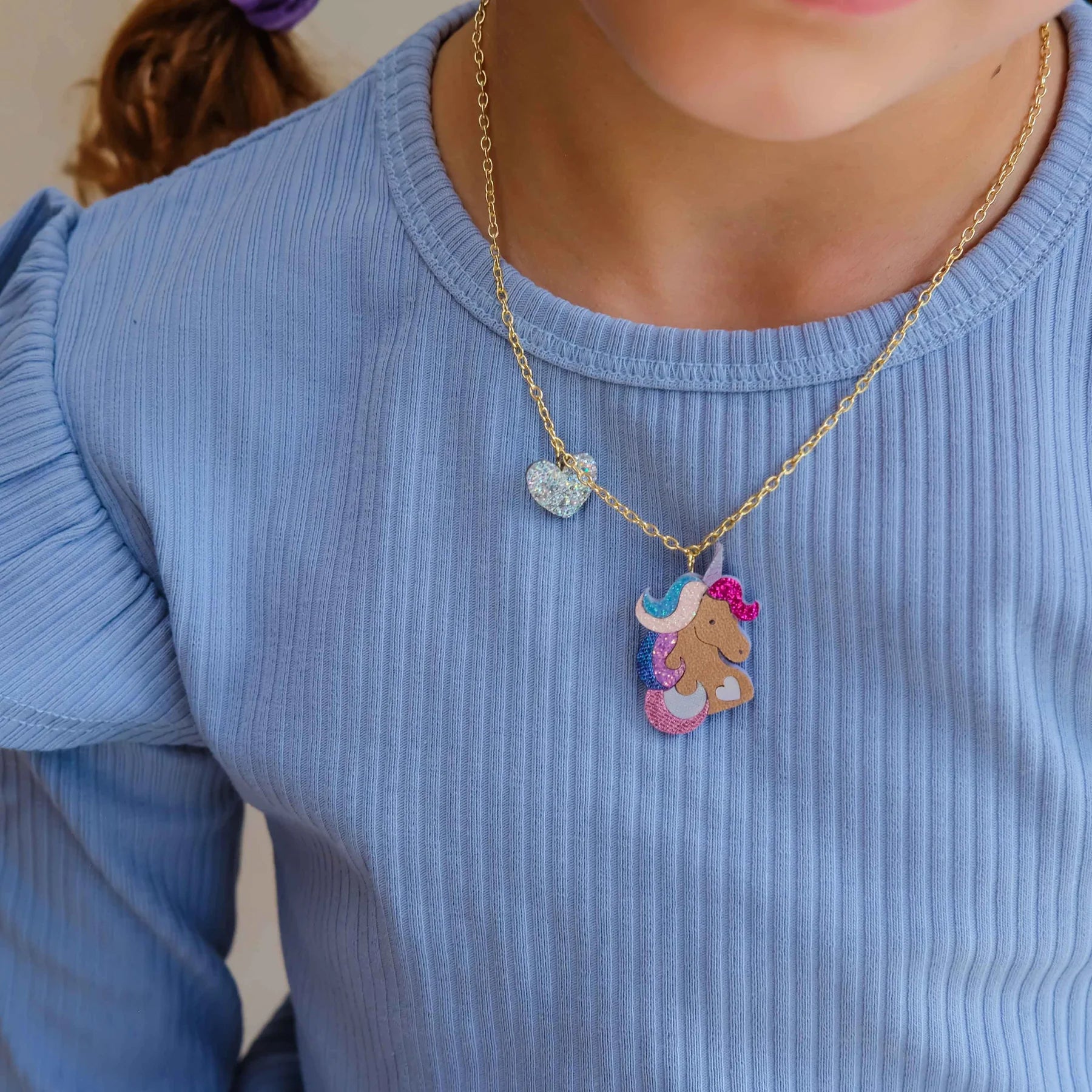 A close-up of a child wearing a gold chain necklace with a colorful unicorn pendant and a small glittery heart charm, paired with a light blue ribbed long-sleeve top.