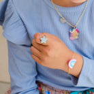 A close-up of a person wearing a light blue ribbed top and jewelry, including a gold necklace with a pastel unicorn pendant, a gold bracelet with a pink and blue rainbow charm, and a glittery star-shaped ring on the finger.