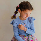 A young girl with brown hair in pigtails wearing a light blue long-sleeve ruffled shirt and a colorful patterned skirt, accessorized with a silver unicorn necklace and matching star ring.