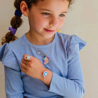 A young girl with brown hair in a ponytail wearing a light blue ribbed long-sleeve top, showcasing a matching jewelry set with a star ring, a unicorn necklace, and a rainbow bracelet.