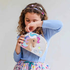 A young girl with curly brown hair wearing a light blue ribbed long-sleeve top and a colorful patterned skirt, holding a small beige unicorn patchwork bag with embroidered details. She is wearing a pastel unicorn headband against a plain light gray background.