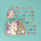 A set of pastel unicorn-themed hair accessories and a matching zip pouch arranged on a light aqua background. The set includes a large unicorn-face pouch, a unicorn keychain, and multiple hair clips and barrettes in pink, blue, yellow, purple, and silver.