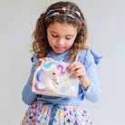 A young girl with curly brown hair wearing a pastel unicorn headband and a blue ribbed top holds a small white shoulder bag decorated with a colorful embroidered unicorn and glittery star details.
